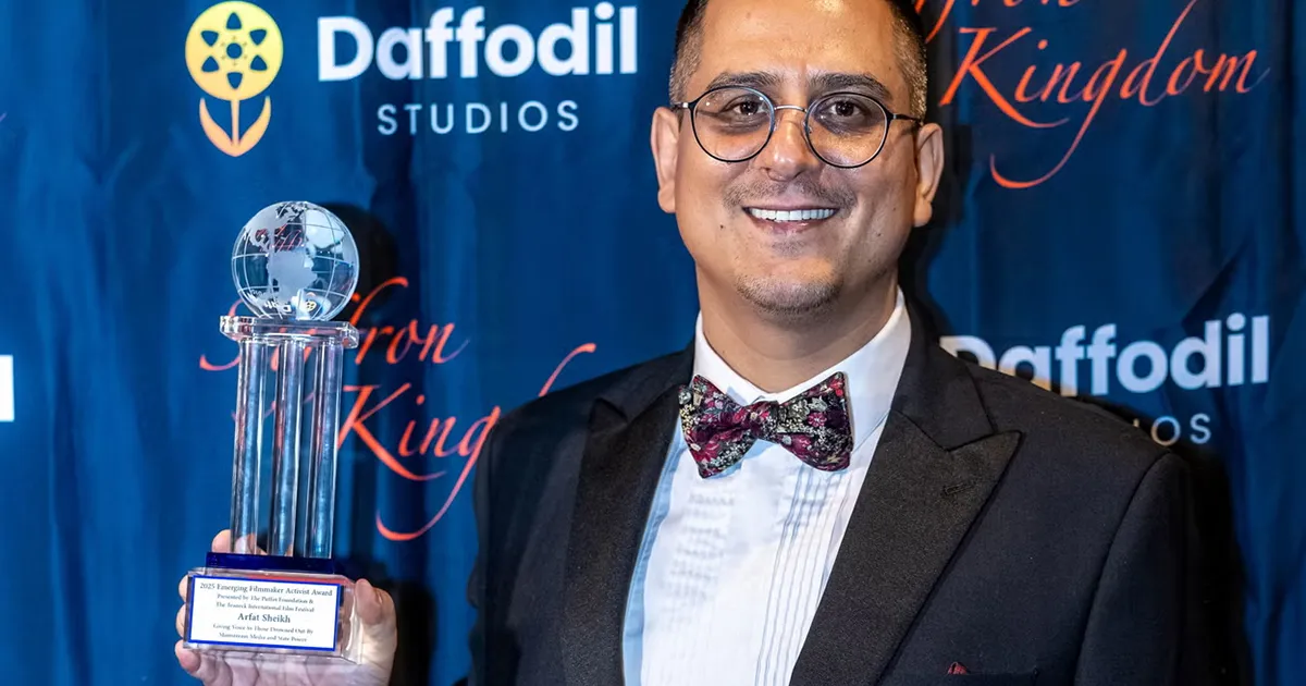A man in a tuxedo holds a trophy before a blue film studio backdrop.