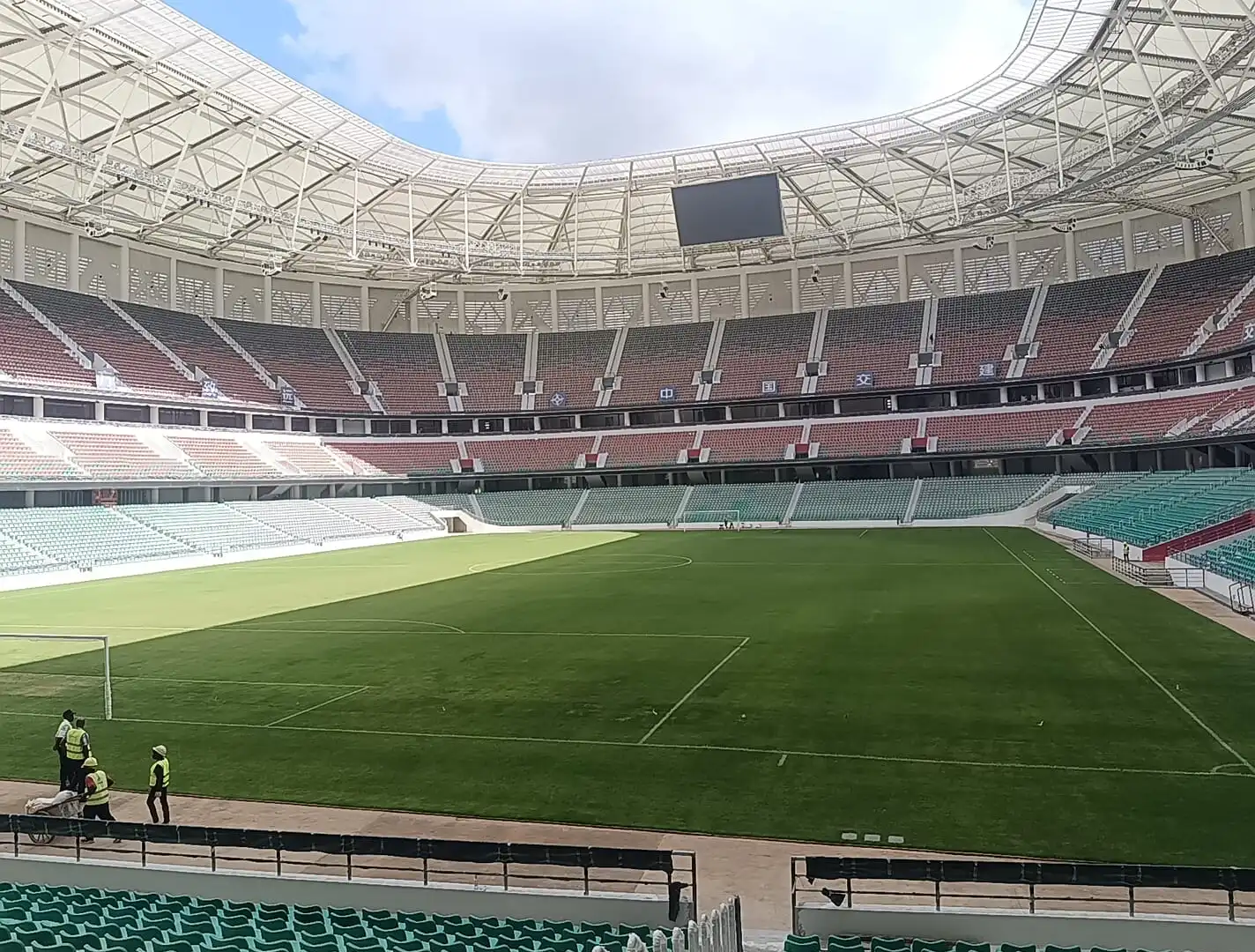Empty football stadium with green pitch, red and teal seating, roof trusses, and workers near the touchline.