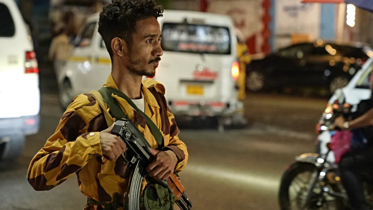 A man in camouflage holds a rifle beside traffic on a city street at night.