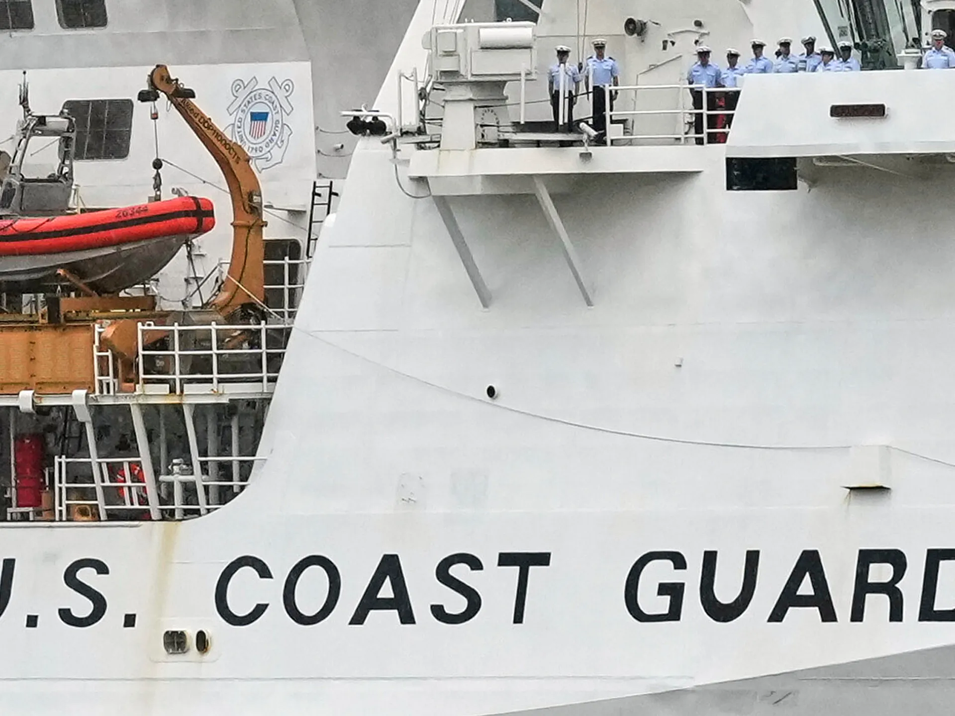 Crew members stand aboard a U.S. Coast Guard vessel beside a suspended red rescue boat.