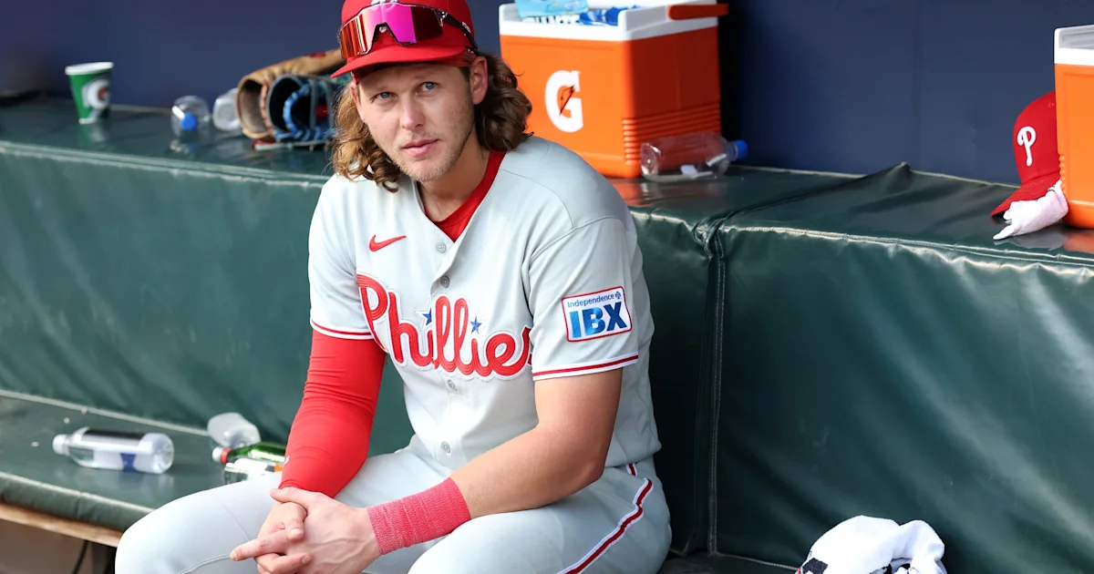 A Phillies player in a gray uniform and red cap sits in the dugout, wearing sunglasses on his cap.