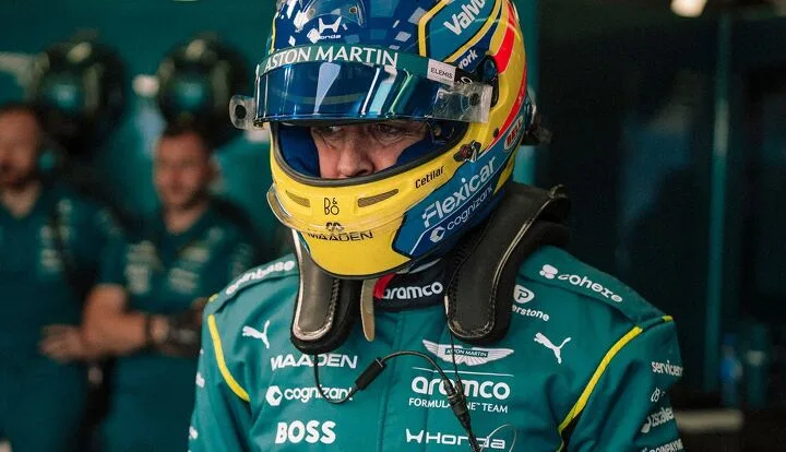 A helmeted driver in a teal Aston Martin racing suit stands among team personnel.