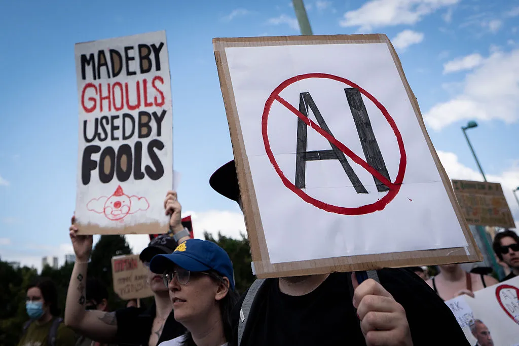 Protesters hold signs, including a crossed-out AI logo and a message criticizing AI use.