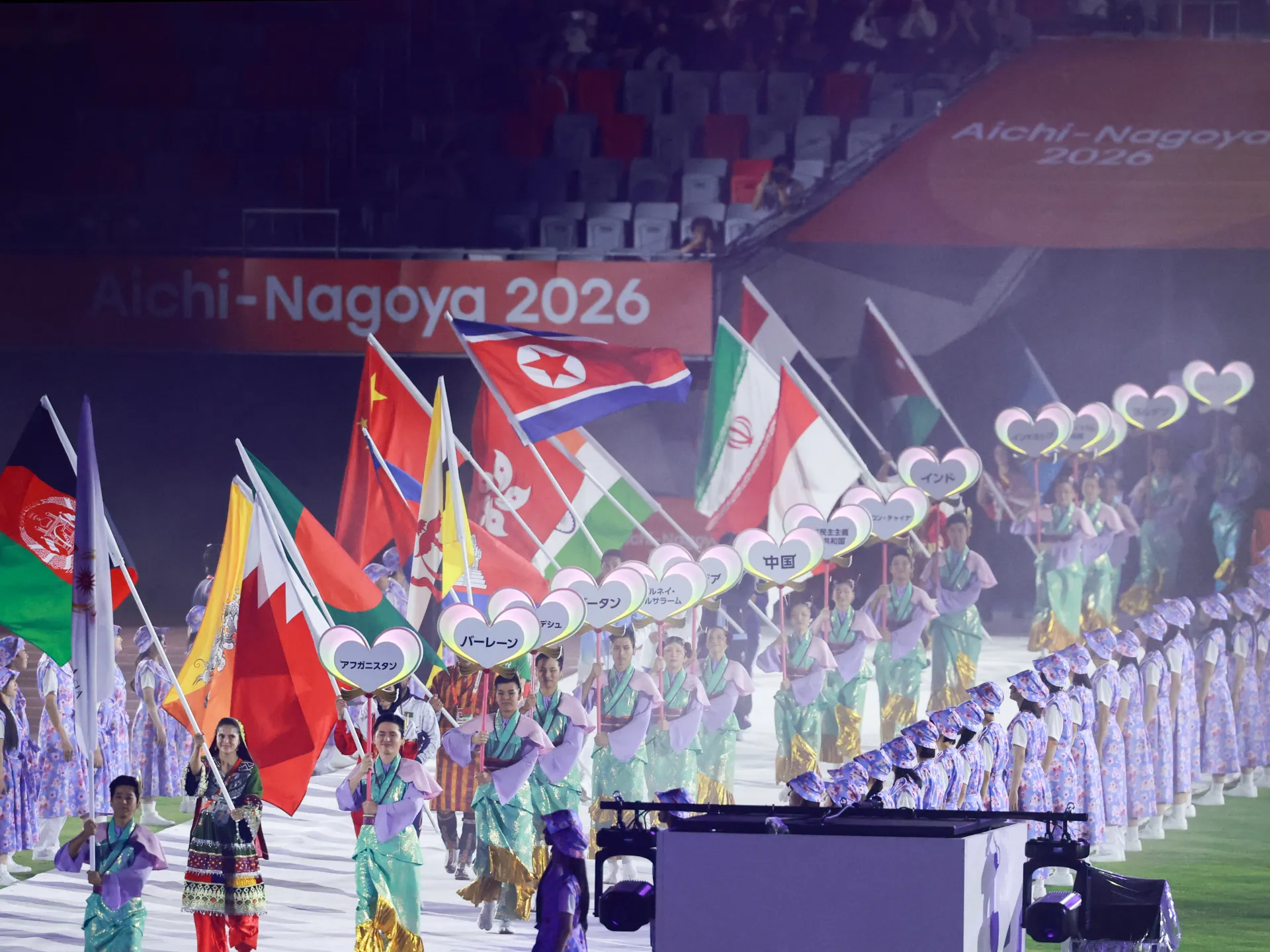 Flag bearers and performers parade during a stadium ceremony, with country signs and an Aichi-Nagoya 2026 banner.