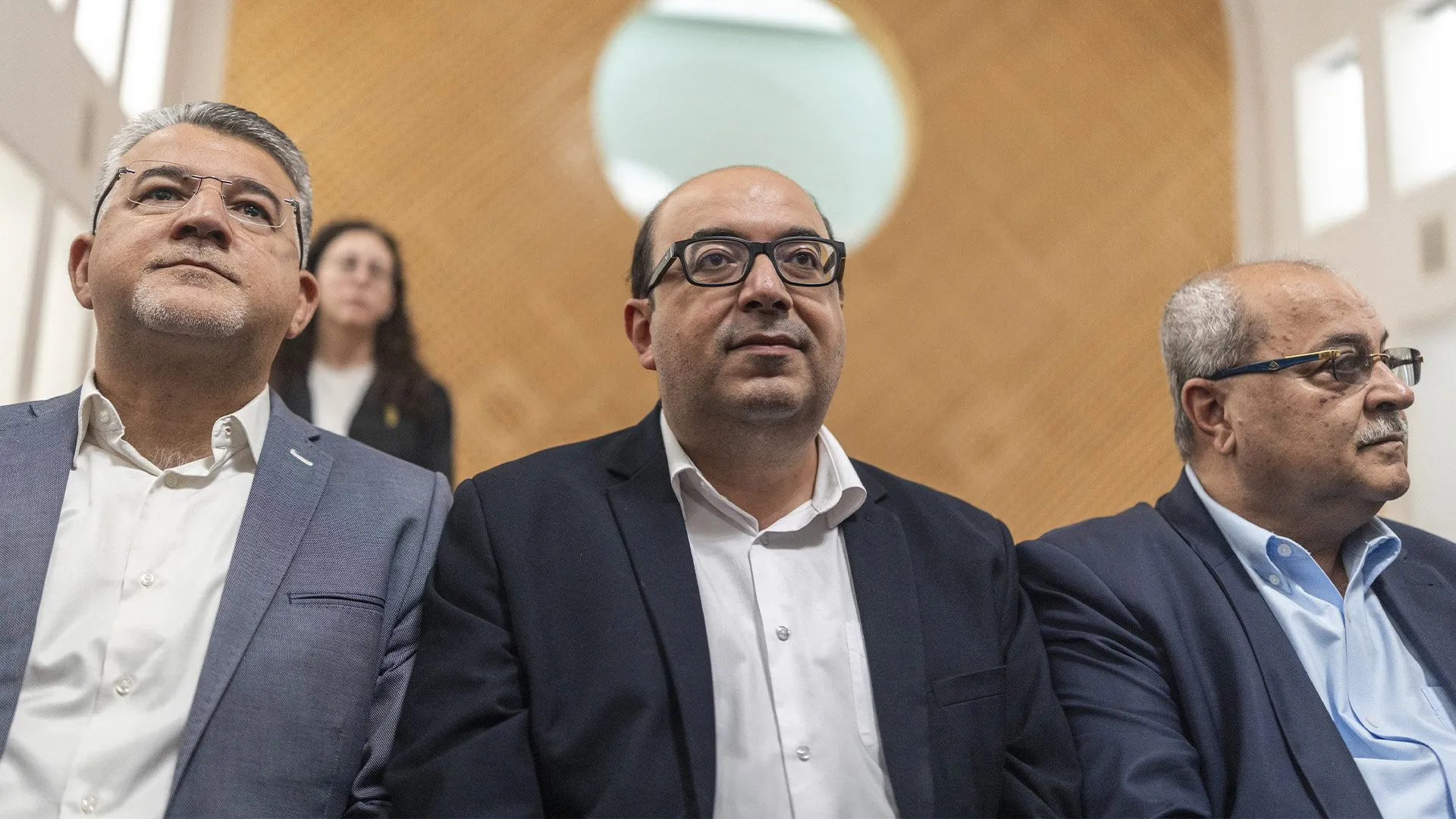 Three men in suits and glasses stand indoors, with a woman visible behind them.
