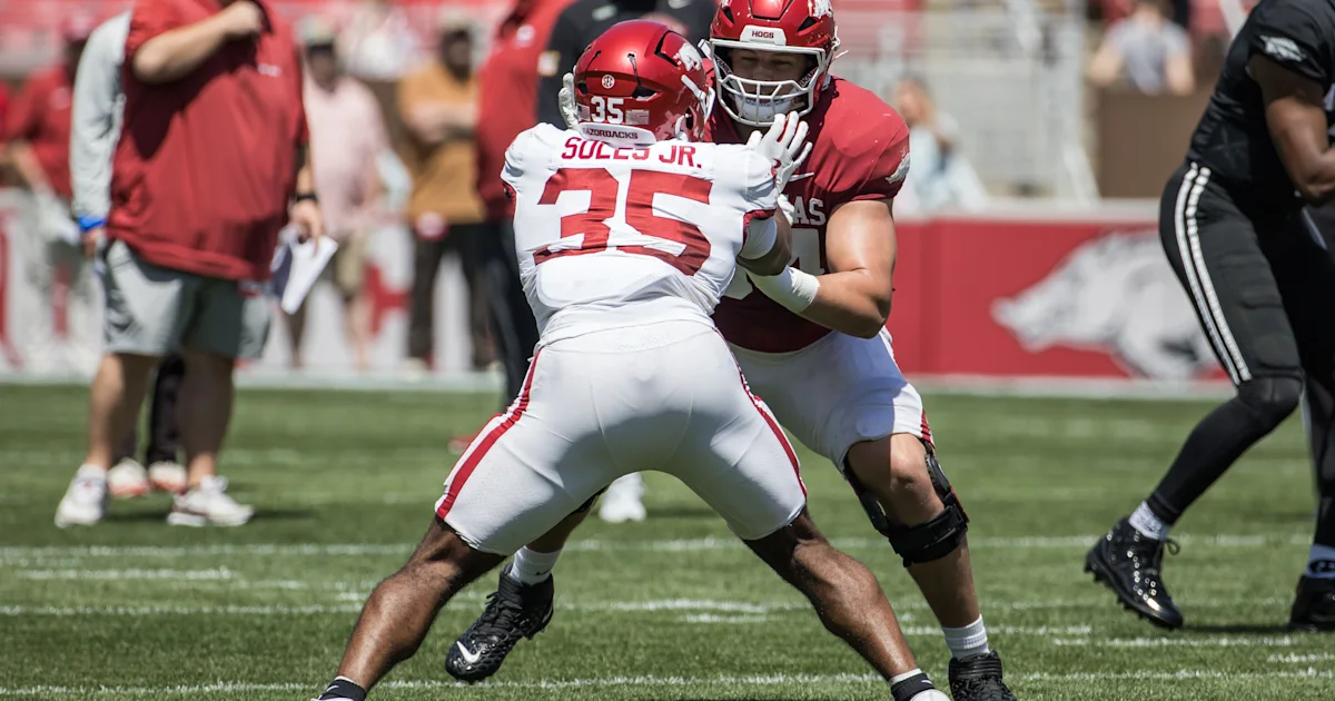 Two Arkansas players in red helmets and practice uniforms engage in a blocking drill on a football field.