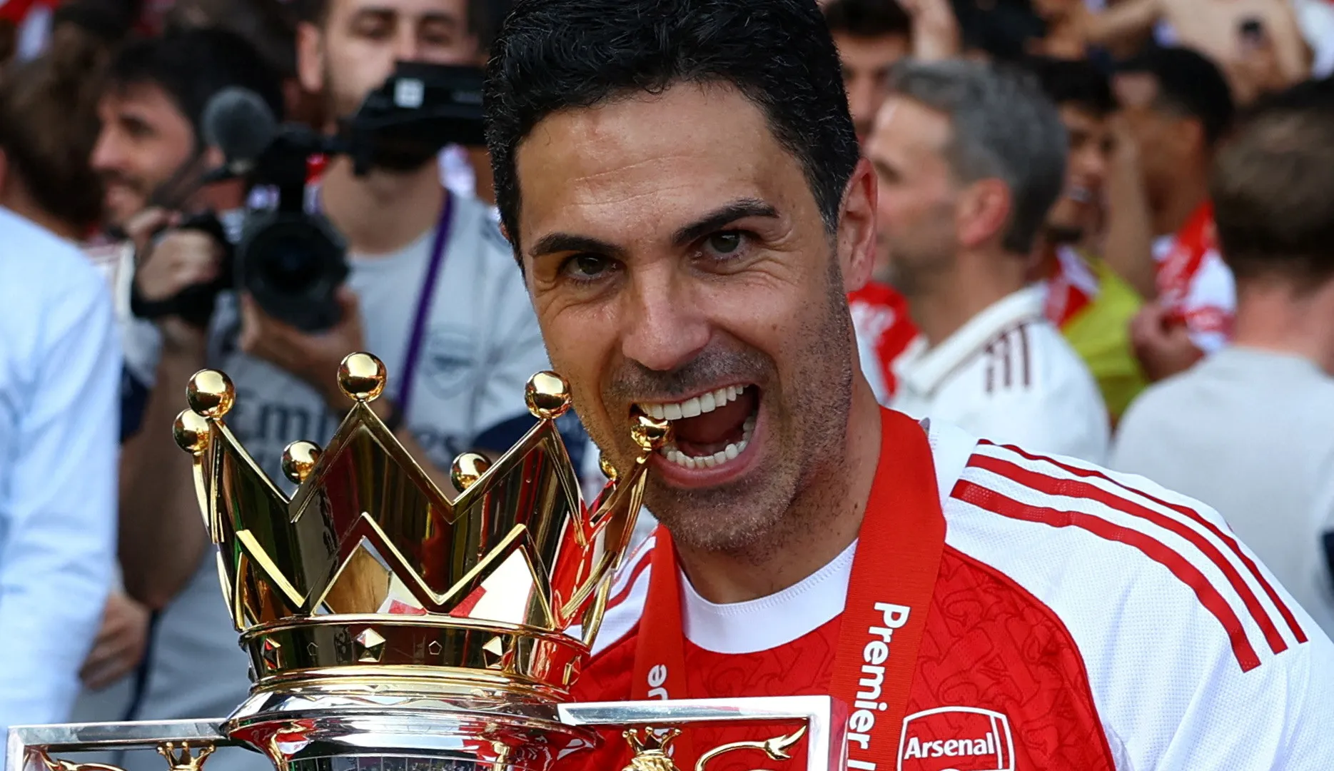 A smiling man in an Arsenal shirt holds a large trophy amid a crowd.