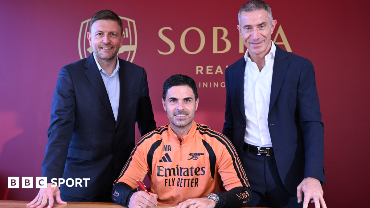 A man in an Arsenal training top signs a document between two men in suits.