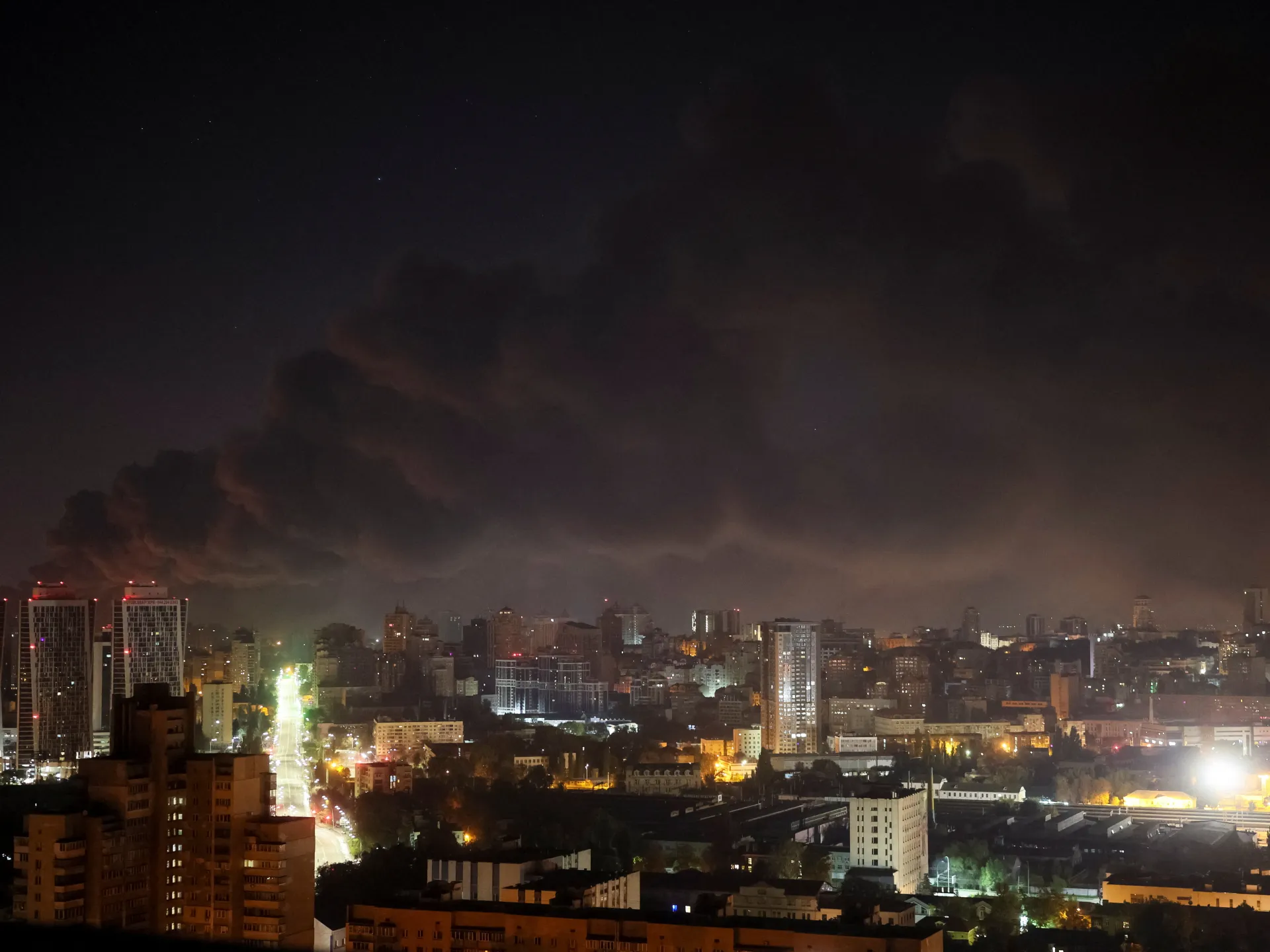 Nighttime city skyline beneath a dark, clouded sky, with illuminated buildings and streets.