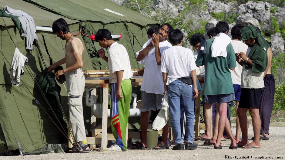 Several men queue beside a green tent, with some washing or changing clothes outdoors.