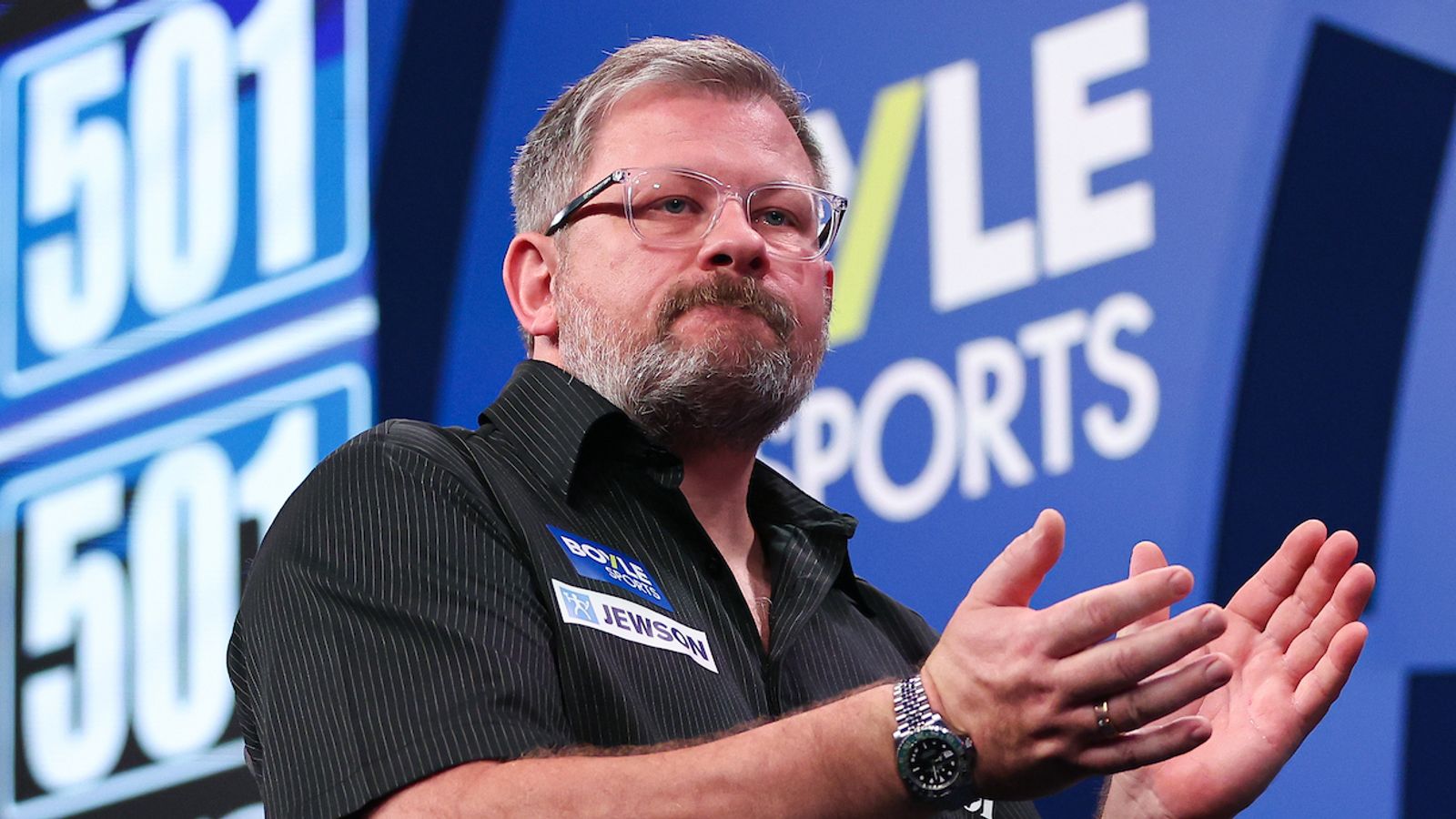 A bearded man in glasses claps onstage in front of blue sports event signage.