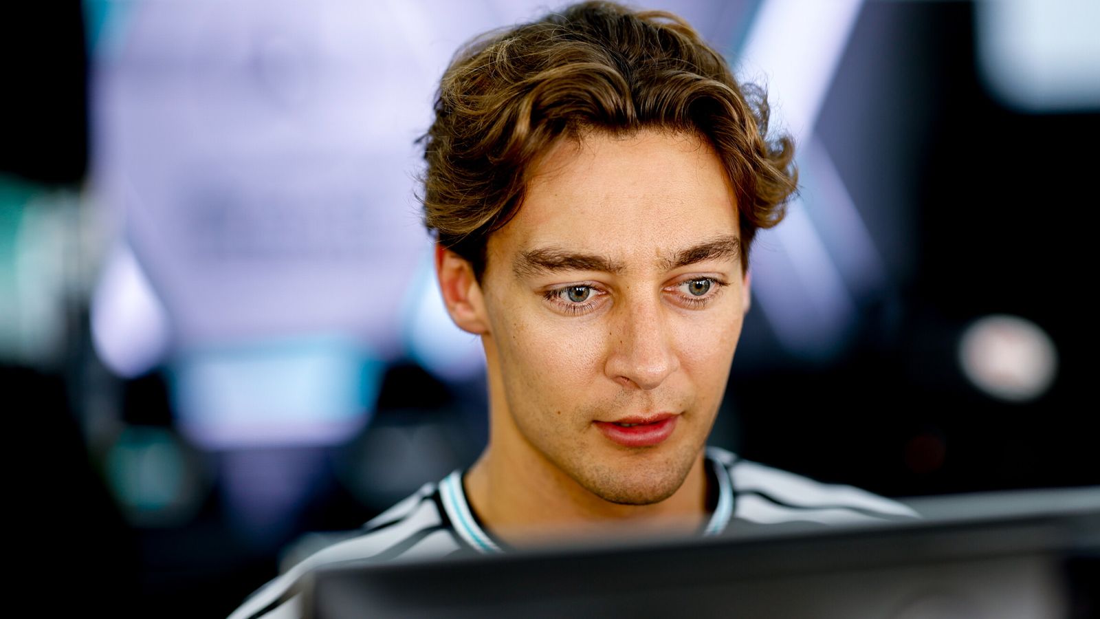 A brown-haired man in a striped shirt looks down toward a monitor in a blurred indoor setting.