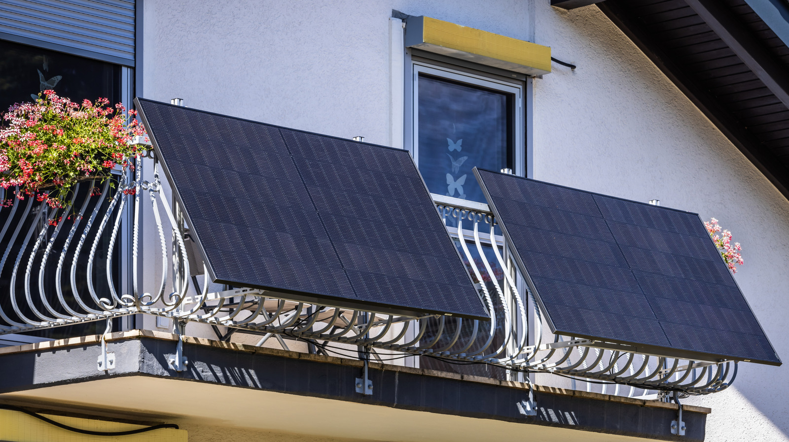 Two dark solar panels mounted on a residential balcony railing beneath windows and flower boxes.