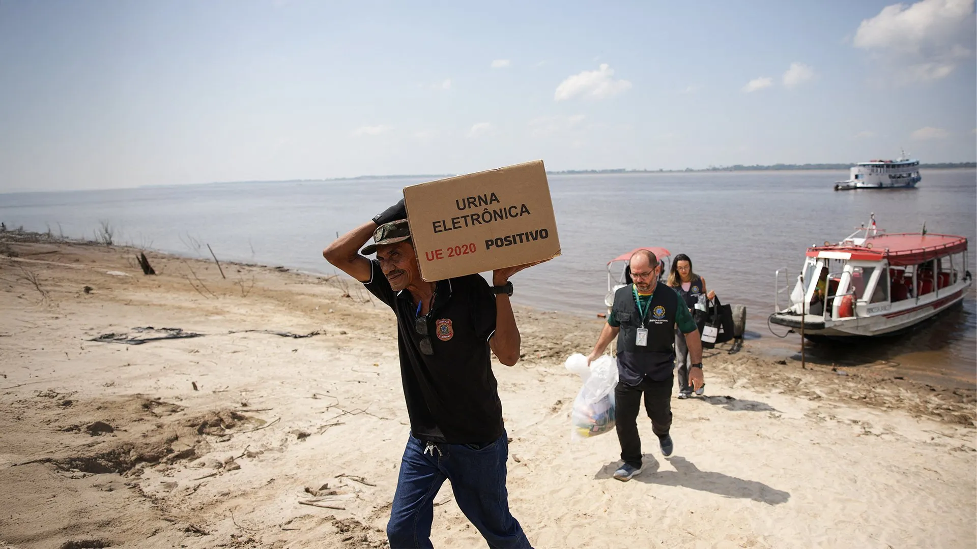 A man carries a box labeled electronic ballot while election workers walk along a sandy riverbank.