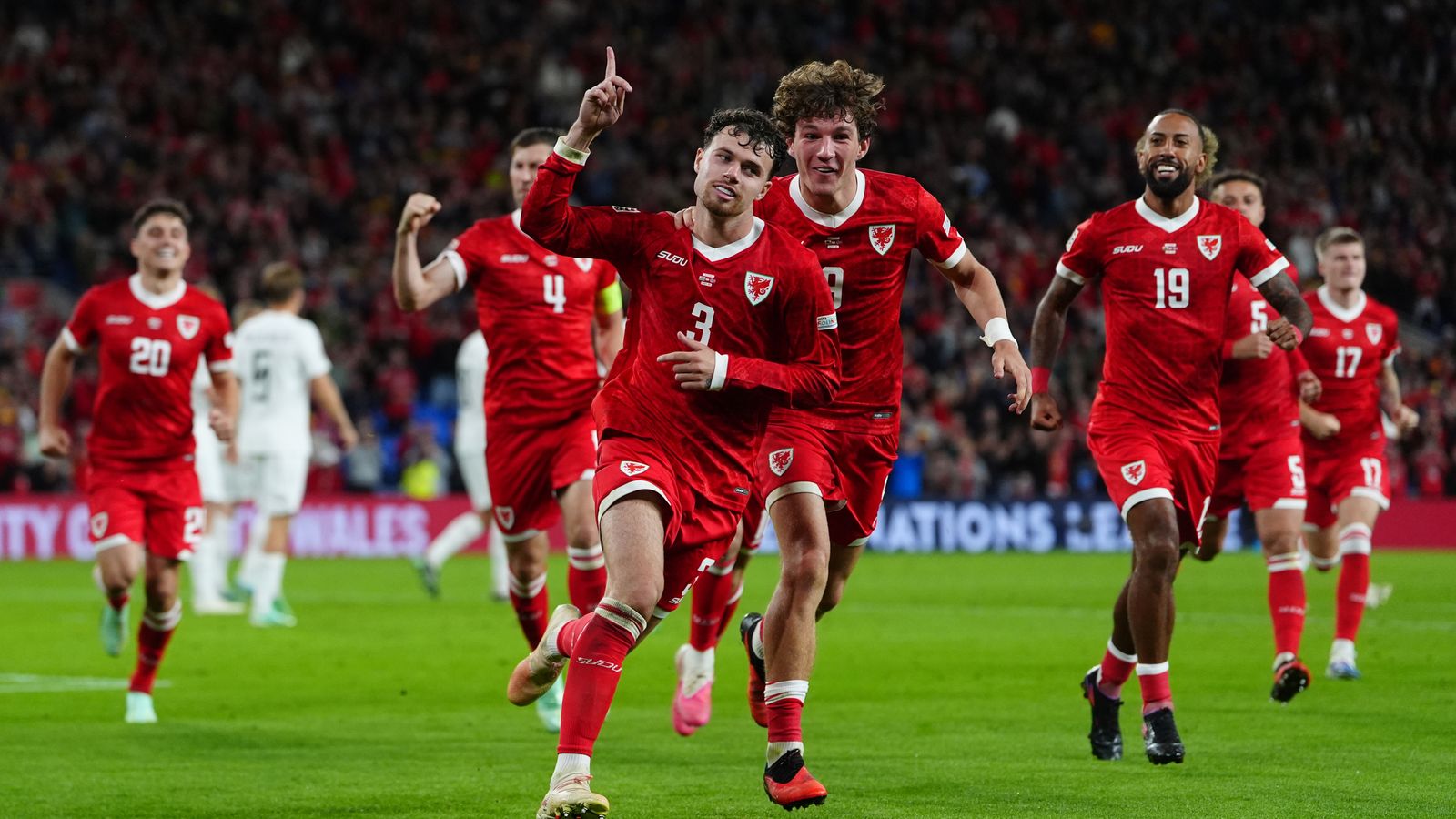 Wales players in red celebrate on the pitch, with one raising a finger as teammates run alongside.