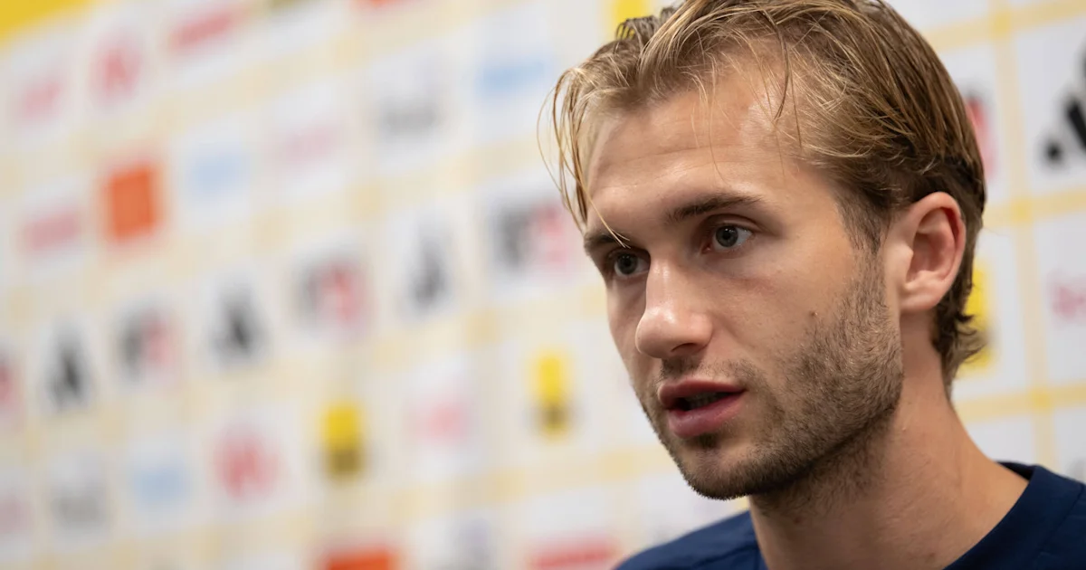 Close-up of a young man with damp, swept-back hair speaking in front of a blurred sponsor backdrop.