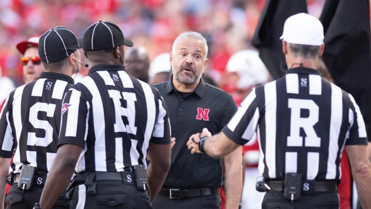 Football coach in a black Nebraska shirt speaks with referees on the field.