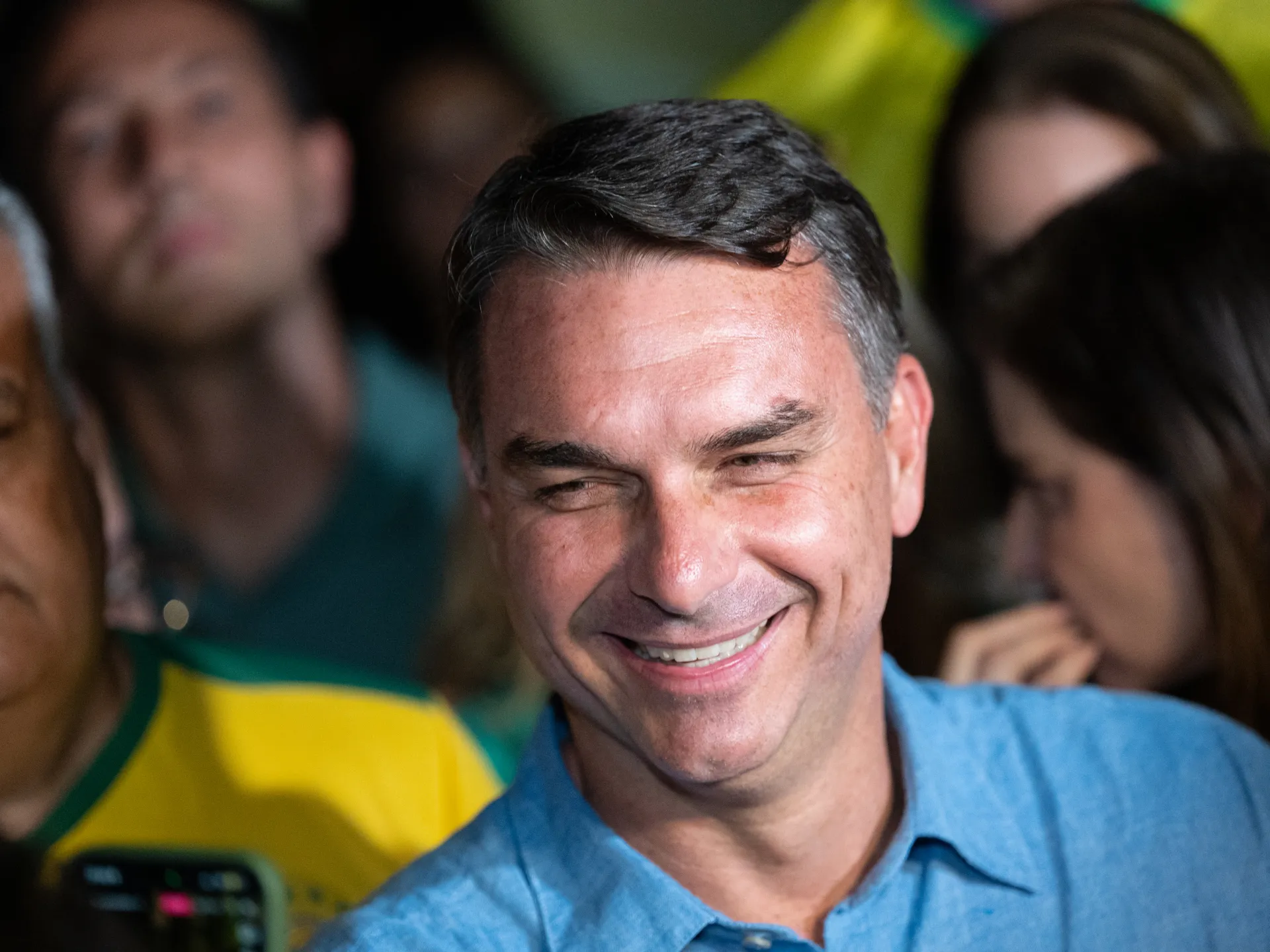 A smiling man in a blue shirt stands among people, with a Brazilian flag visible behind him.