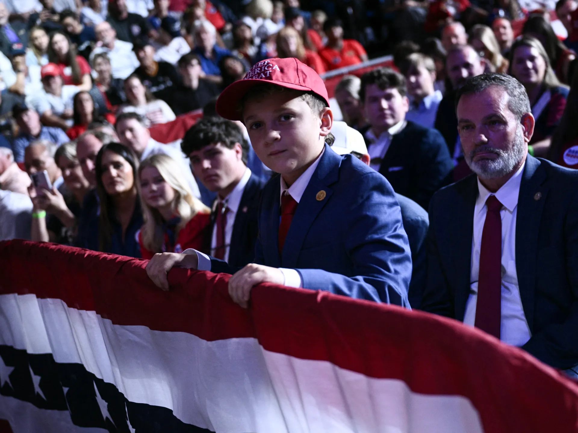 A boy in a red cap and blue suit stands behind a large American flag at a crowded rally.