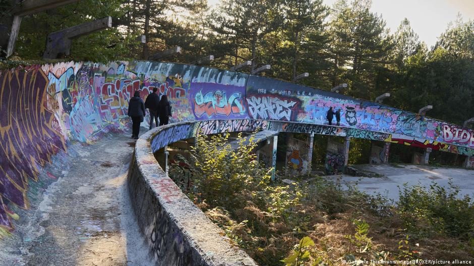 People walk along a curved path beside a graffiti-covered concrete structure among trees.