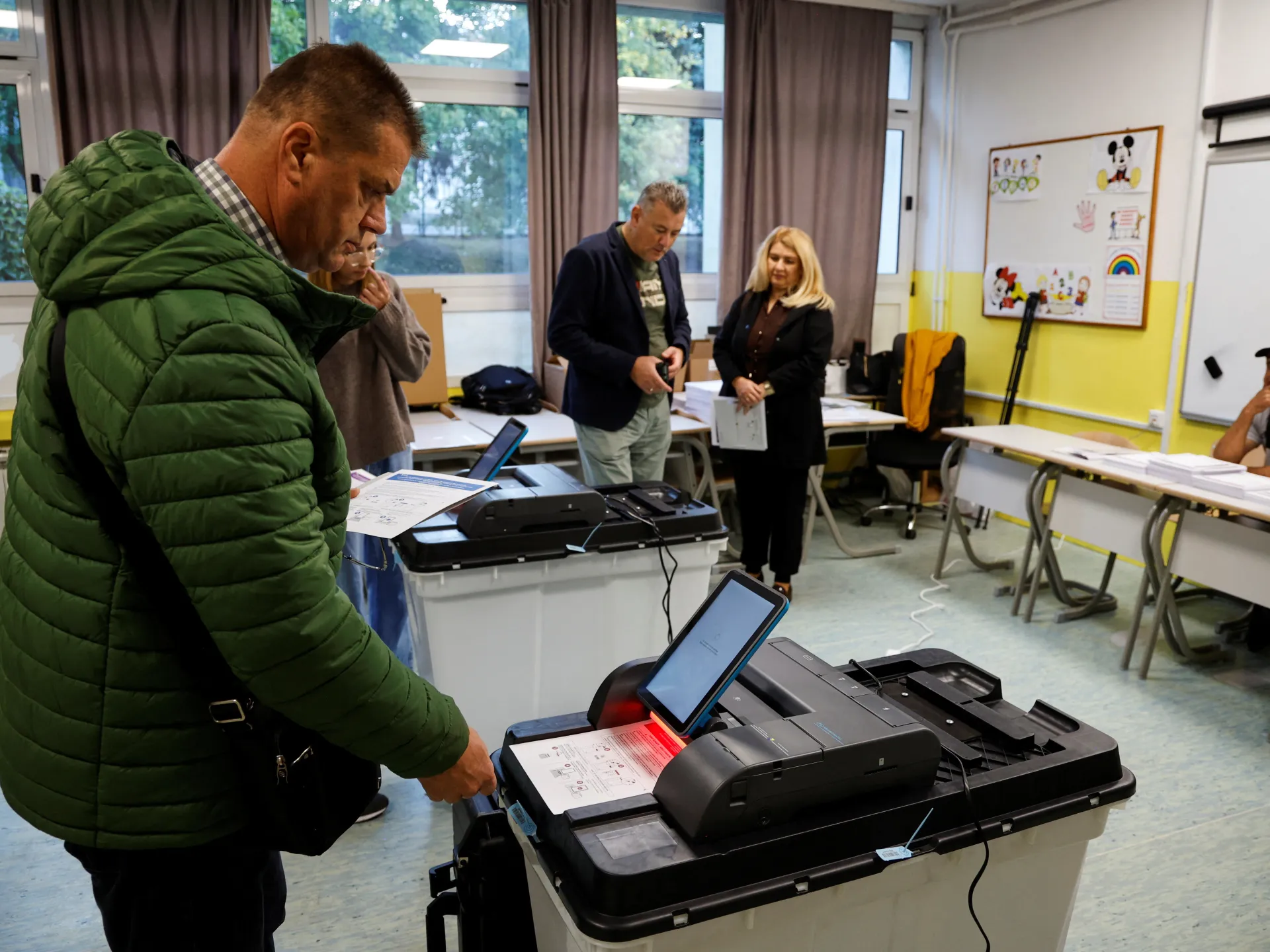 A man in a green jacket uses a ballot scanning machine in a classroom polling station.