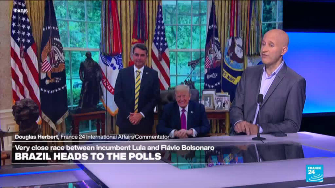 A France 24 commentator speaks beside a screen showing Donald Trump seated with another man among flags.