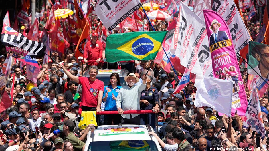 Crowd surrounds a campaign vehicle as a man waves, with Brazilian flags and political banners overhead.