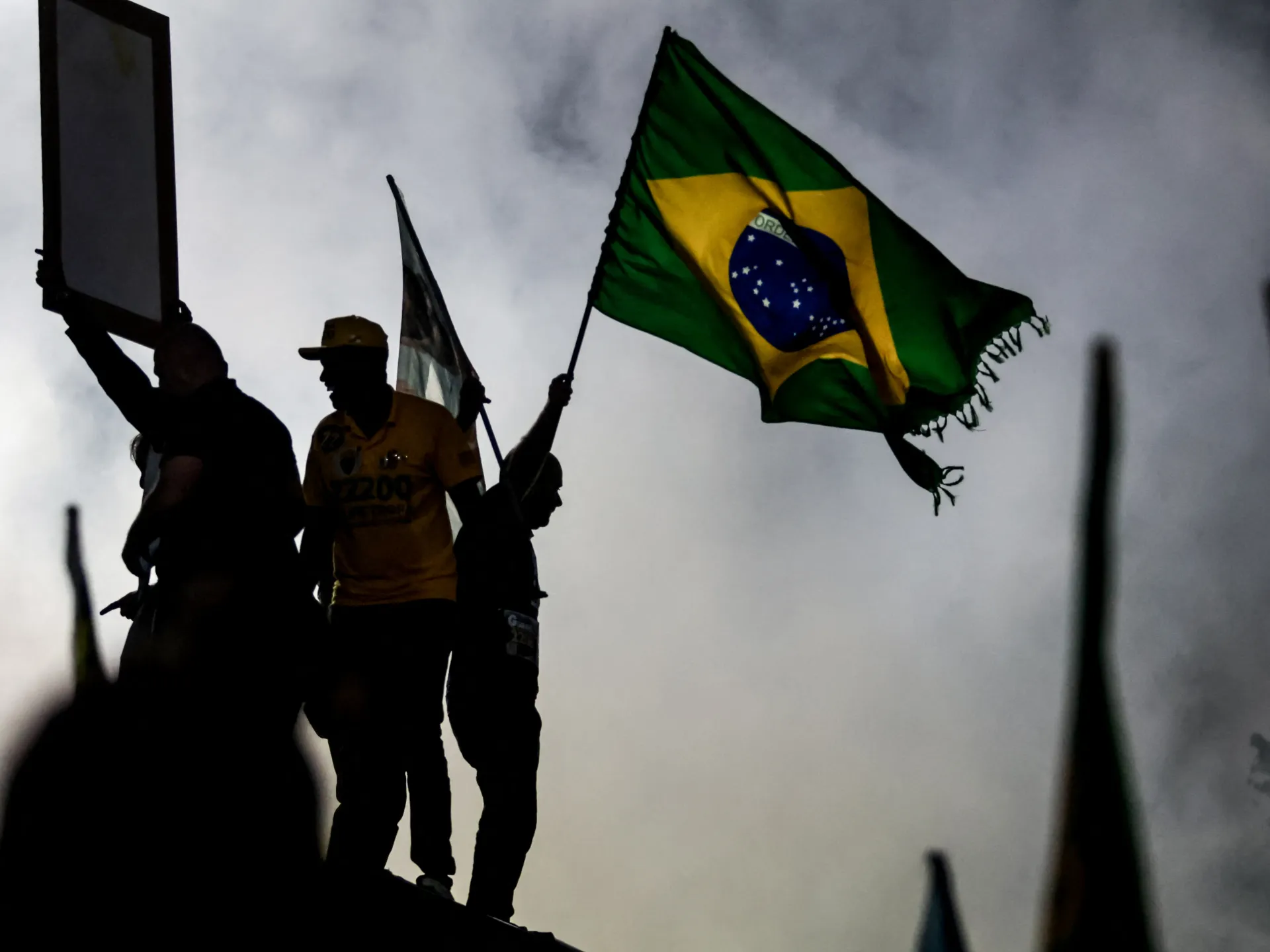 Silhouetted people stand beneath a Brazilian flag, with one person holding a rectangular sign aloft.