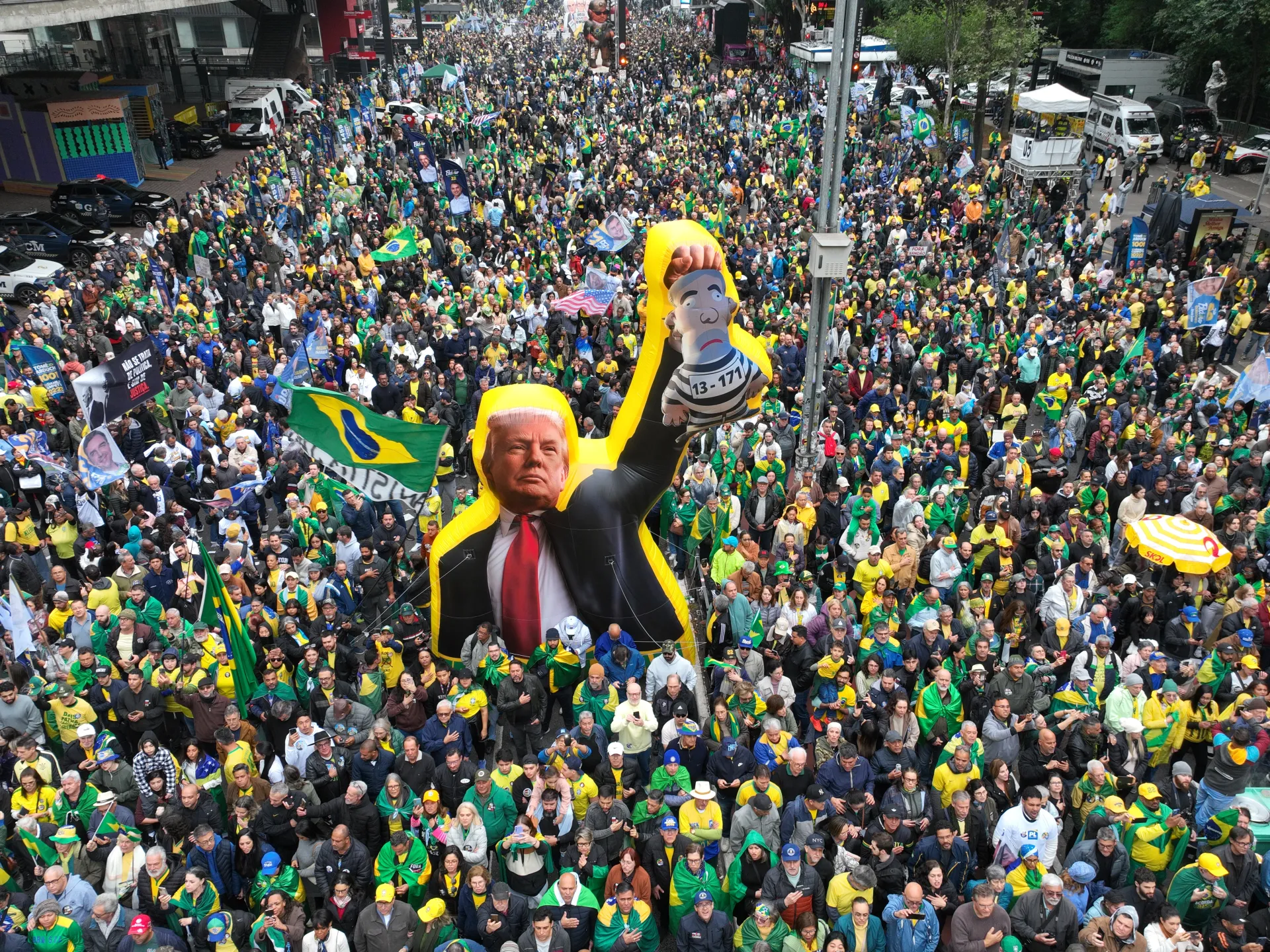 A dense crowd in Brazilian colors gathers around large caricature balloons of Donald Trump and a prisoner.