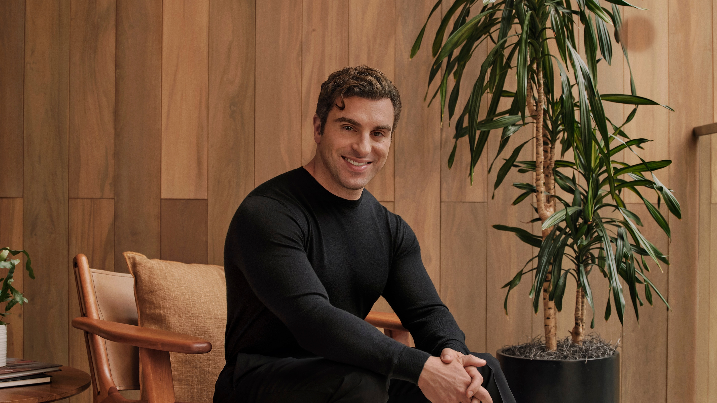 A smiling man in a black sweater sits beside a potted plant against a wood-paneled wall.