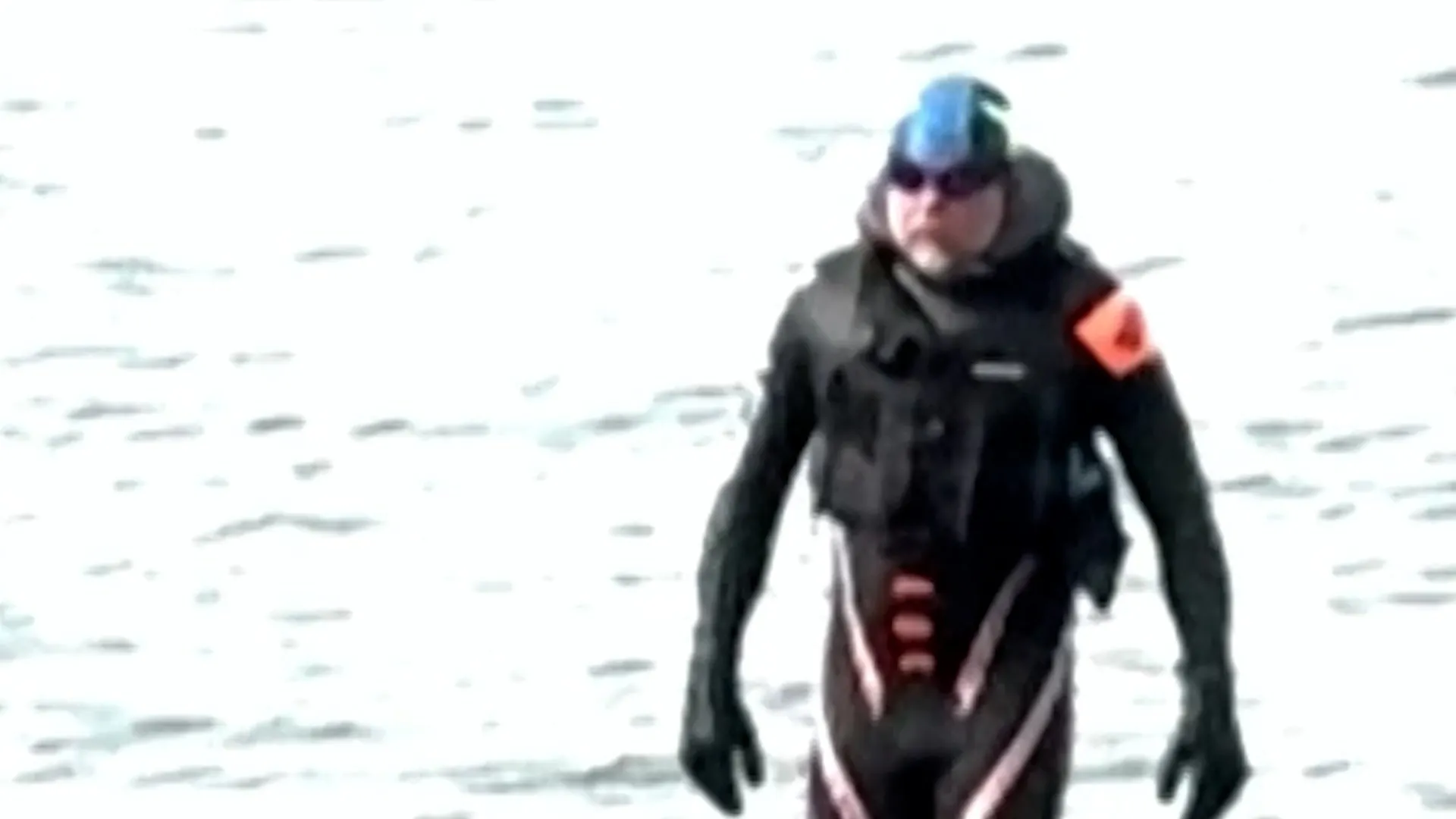 A person in dark cold-weather clothing and sunglasses walks across a snowy landscape.