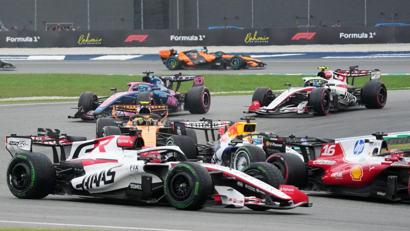 Several Formula One cars race closely together on a circuit, with Bahrain Grand Prix signage in the background.