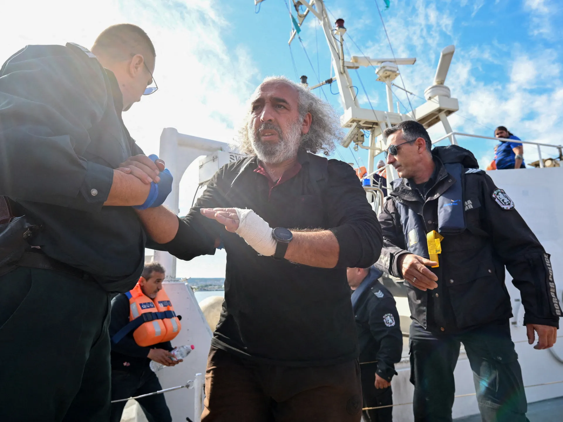 A bandaged man speaks with uniformed personnel aboard a vessel under a bright, partly cloudy sky.