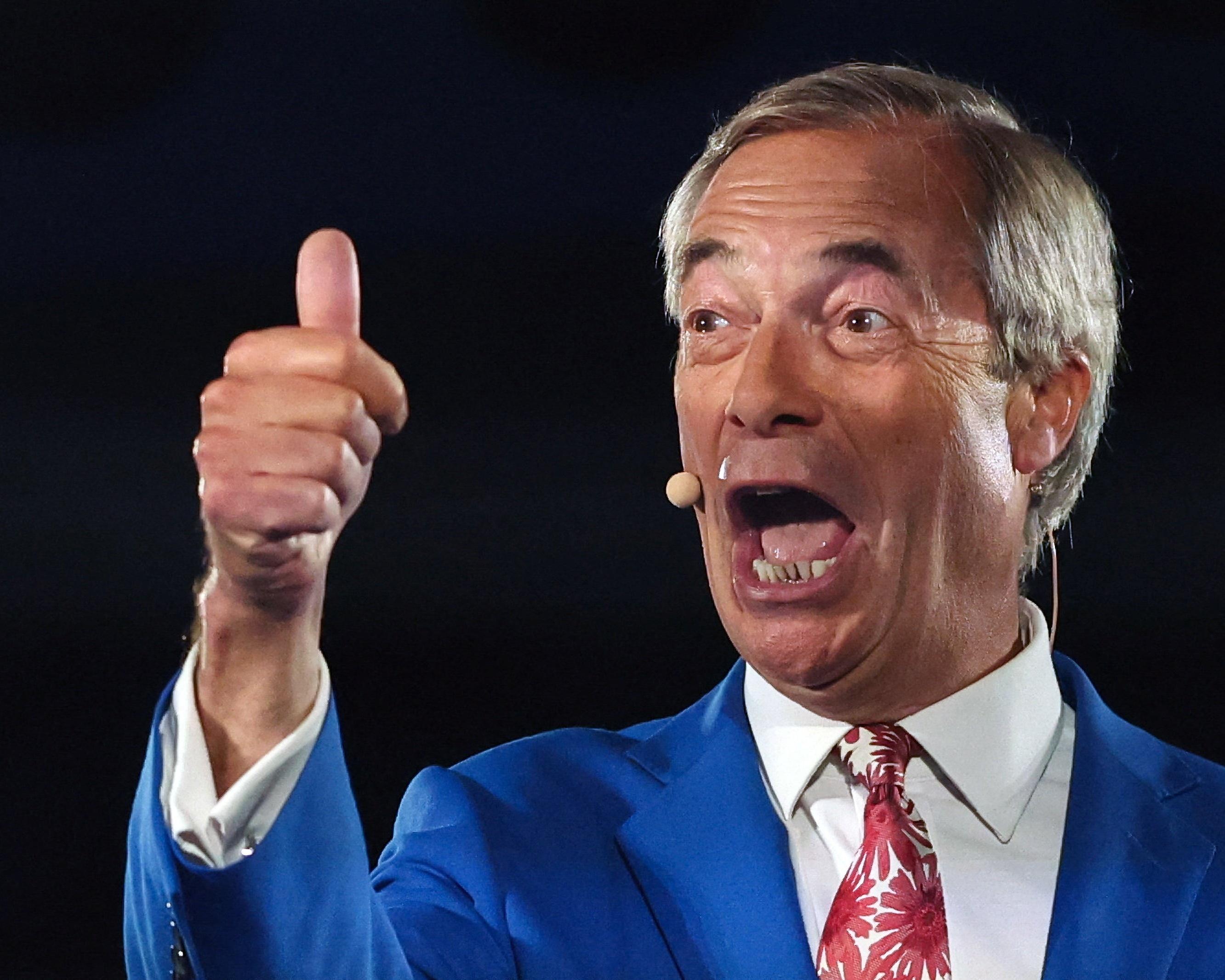 A gray-haired man in a blue suit speaks into a headset microphone while giving a thumbs-up.