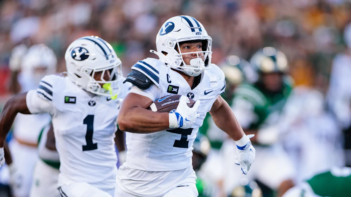 BYU player in white uniform carries the football as a teammate follows during a game.