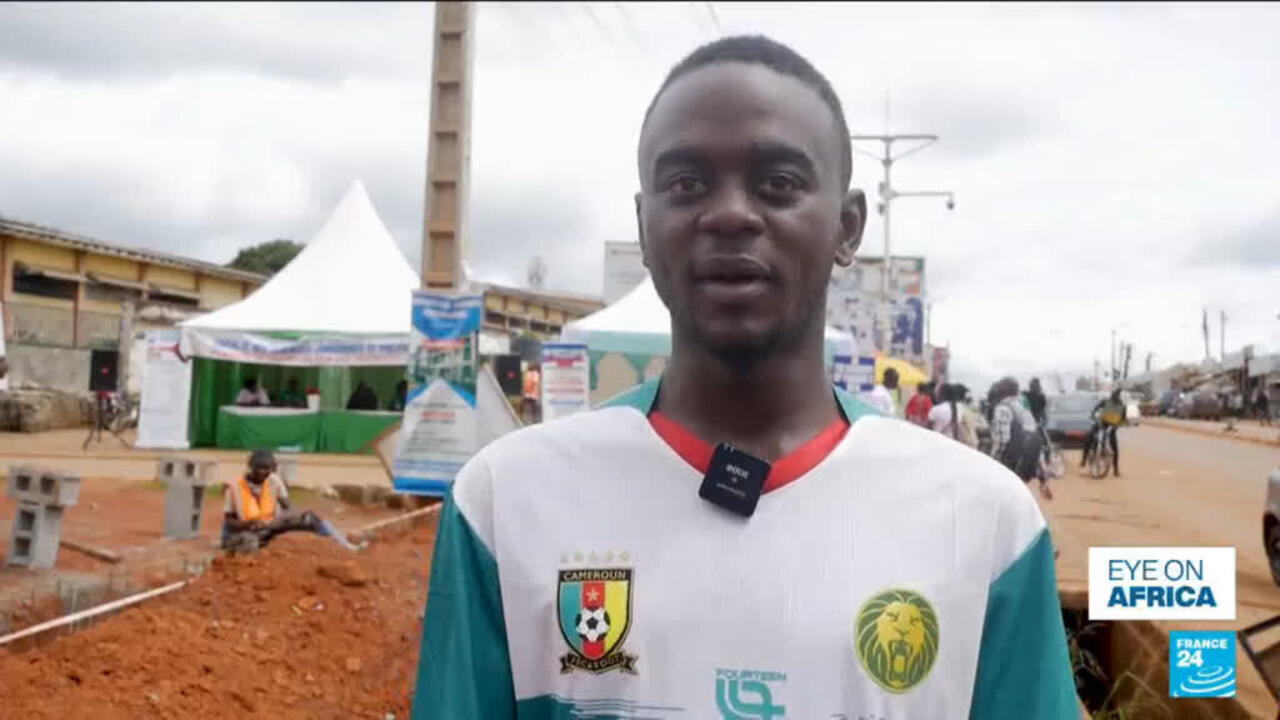 A man in a Cameroon football jersey stands outdoors near tents and a roadside construction area.