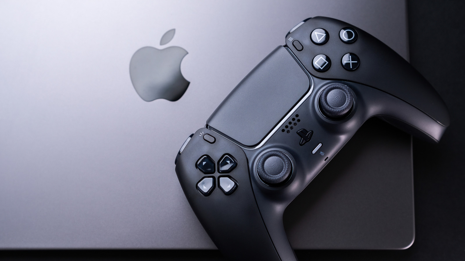 A black game controller rests diagonally across a silver laptop displaying an Apple logo.