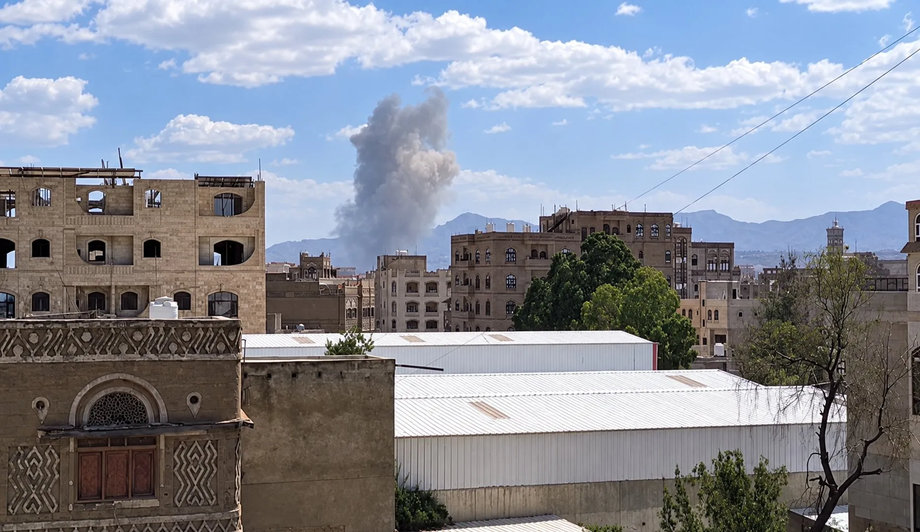 A plume of gray smoke rises behind city buildings beneath a blue, cloud-filled sky.