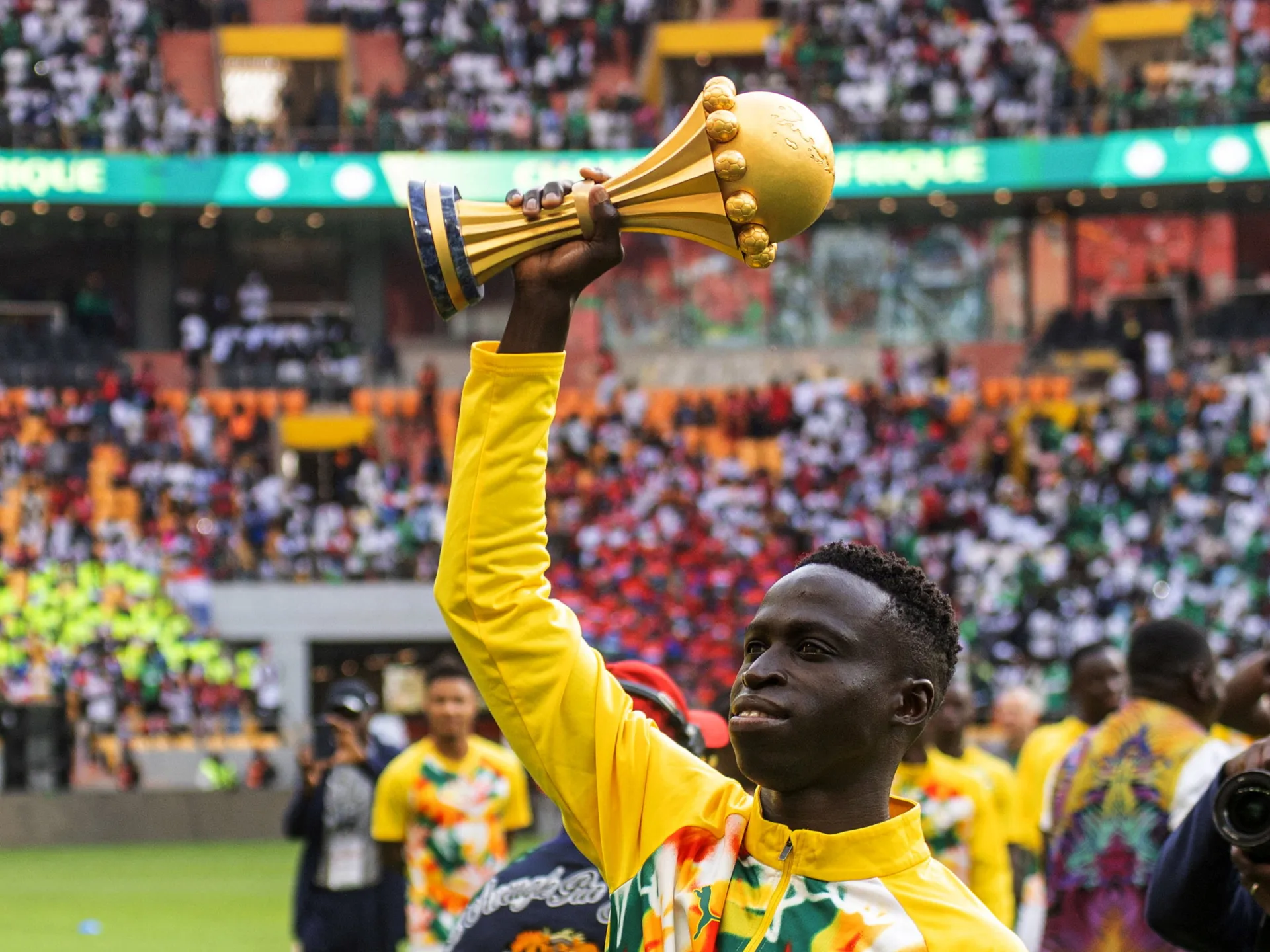 A man in a yellow tracksuit raises a gold football trophy above a crowded stadium.