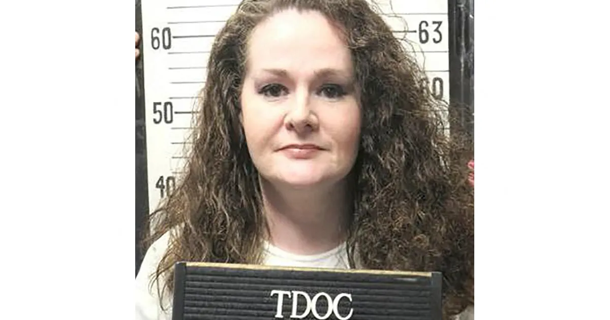 A woman with long curly hair stands before a height chart, holding a black TDOC placard.