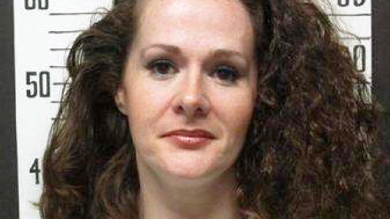 A woman with long brown hair faces the camera in front of a height chart.