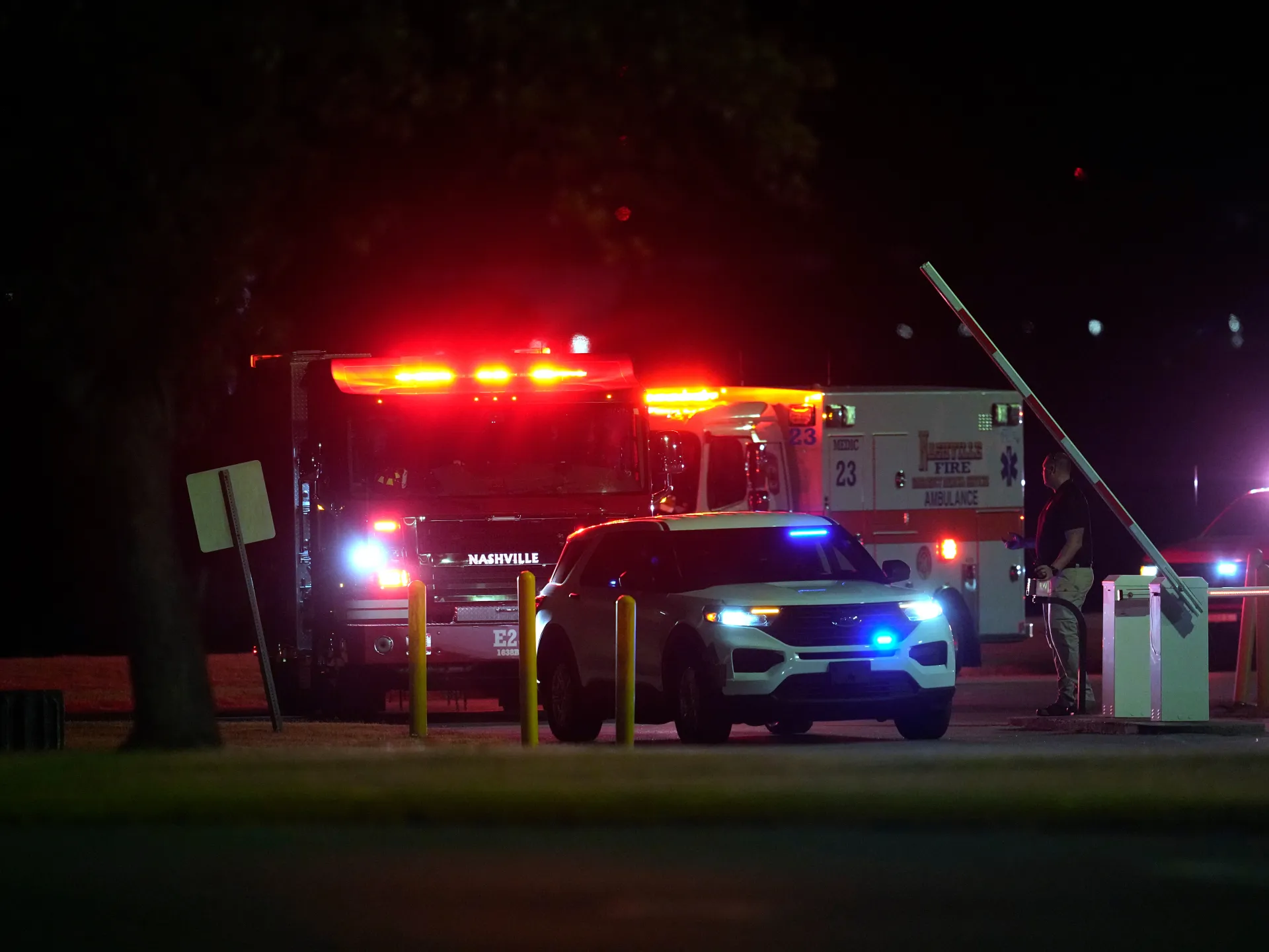 Emergency vehicles with flashing lights gather at night near a raised barrier and a uniformed person.