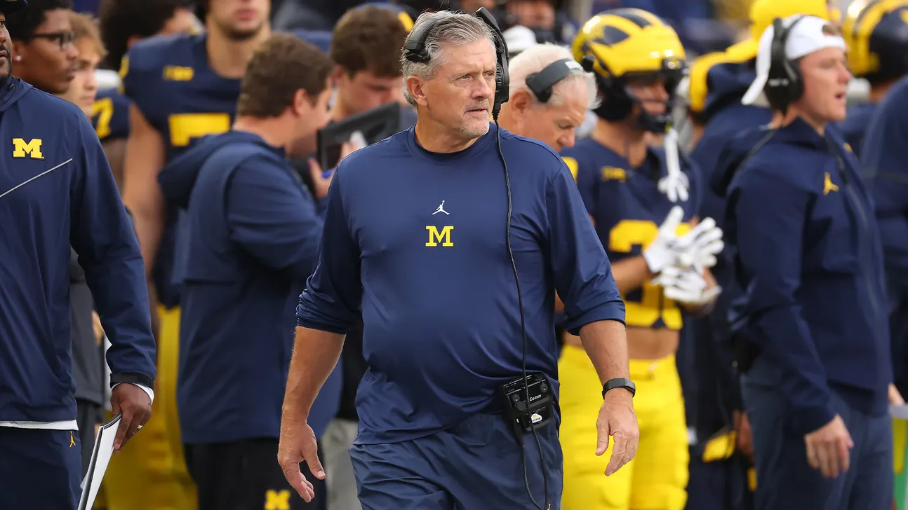 A gray-haired coach in navy Michigan gear walks along the sideline wearing a headset.