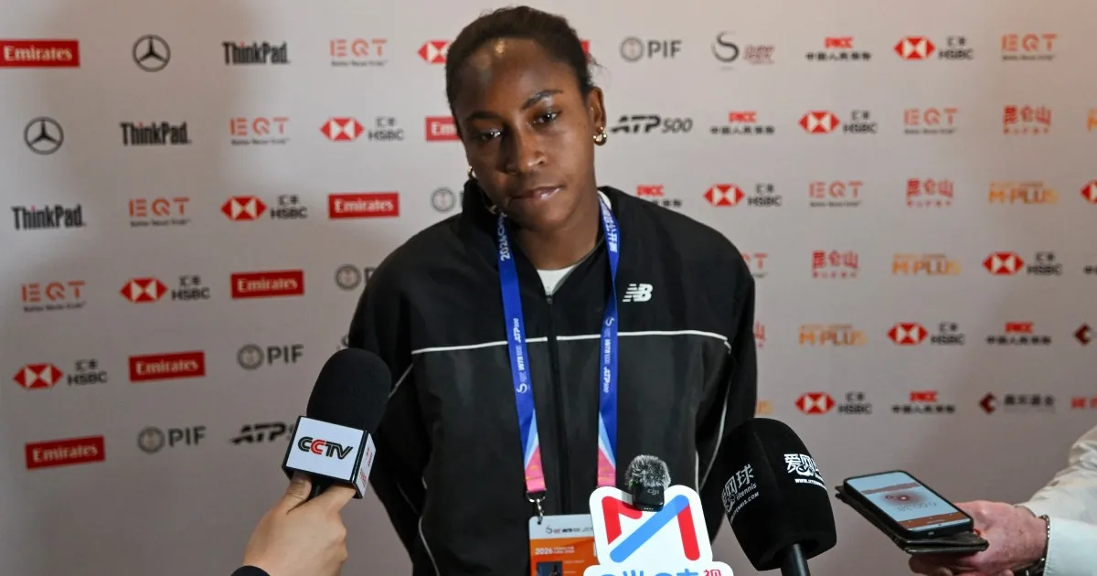 A tennis player in a black jacket speaks to reporters as microphones and a phone are held toward her.