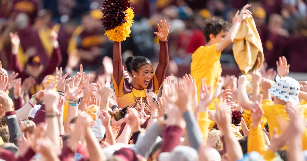 A Minnesota cheerleader raises her arms above a cheering crowd as another holds up a gold cloth.
