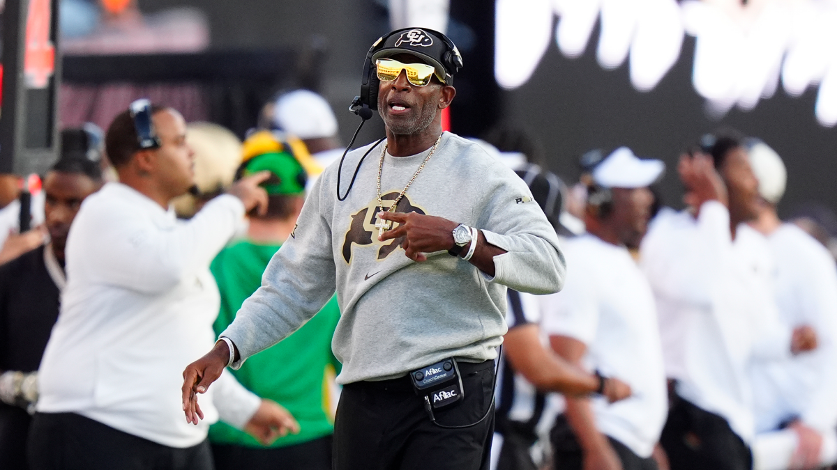A coach in a Colorado sweatshirt gestures on the sideline while wearing a headset and cap.