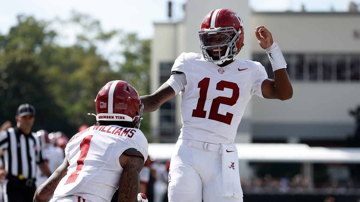 Alabama players in white uniforms celebrate on the field, with number 12 raising his arm beside a teammate.
