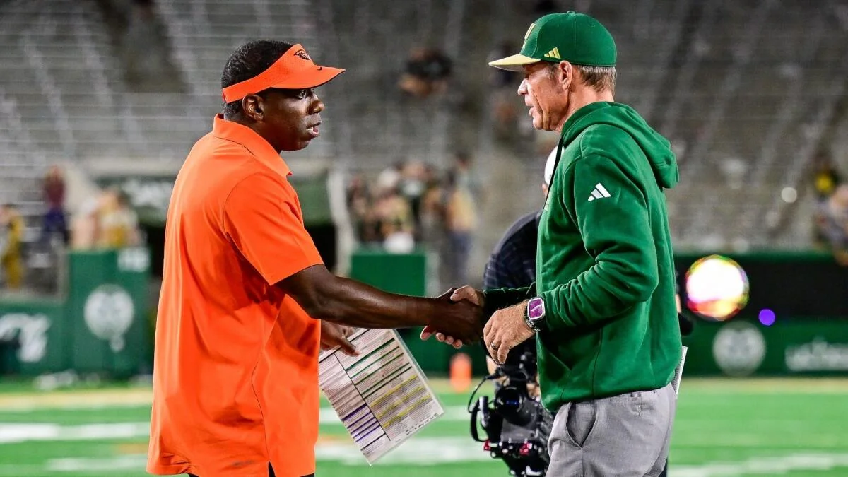 A man in an orange shirt shakes hands with a man wearing a green hoodie and cap on a football field.