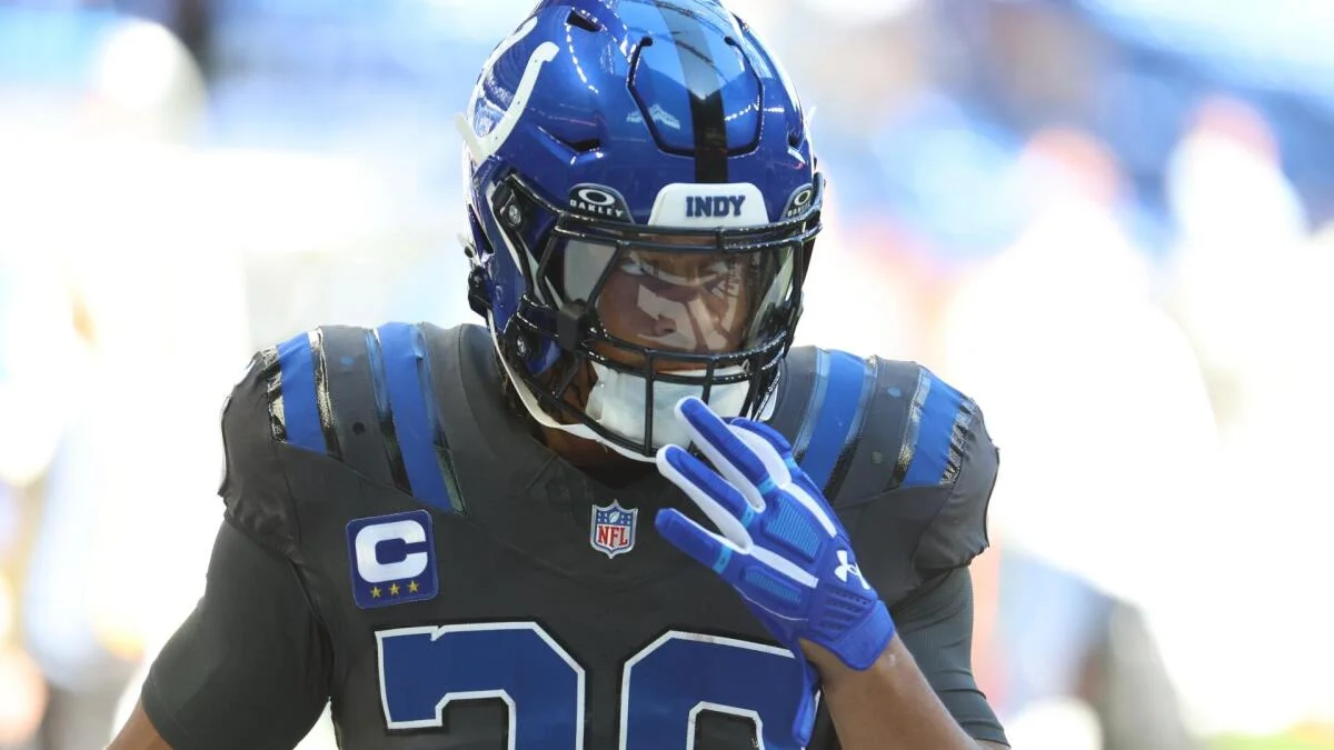 Football player in a blue helmet and dark jersey raises a gloved hand near his face.