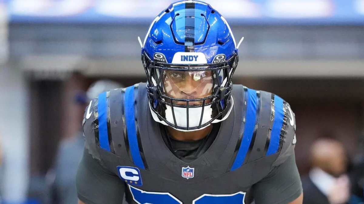 Football player in a blue helmet and gray uniform crouches, looking forward on the field.